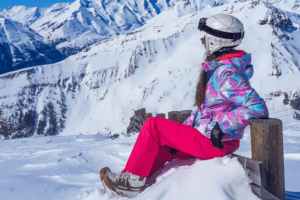 Mädchen mit bunter Skijacke sitzt im Schnee und schaut in die Ferne